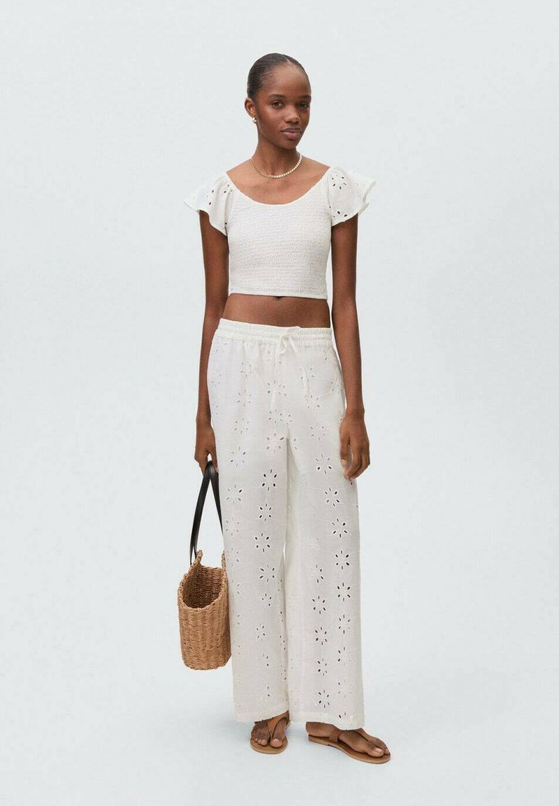 Top bianco a maniche voluminose, abbinato a pantaloni bianchi a gamba larga con ritagli floreali. Il modello tiene una borsa intrecciata e indossa sandali.