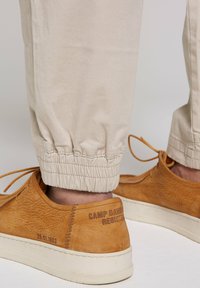 Pantalons beiges fuselés avec des poignets élastiques, associés à des chaussures en cuir texturé marron à semelle claire et détails de talon signés.