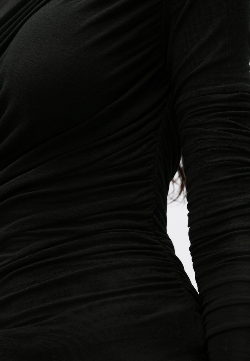 Sort, tætsiddende beklædningsgenstand med teksturerede, rynkede stofdetaljer langs siderne, der fremhæver formerne og skaber en strømlinet silhuet.