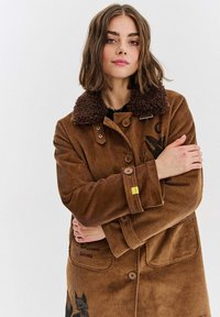 Brązowy płaszcz z korduroju z kołnierzem z futra sztucznego, z guzikami, dwoma przednimi kieszeniami i haftowanymi detalami. Teksturowany, ciepły design.
