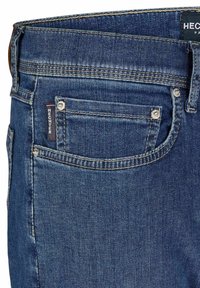 Jean en denim bleu foncé, de coupe classique, avec des coutures visibles, une petite poche avant avec une étiquette de logo et des accents en métal.