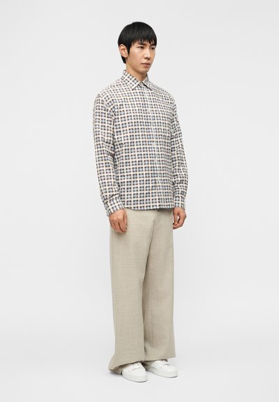 Chemise à manches longues à carreaux blanc, bleu et marron avec un col à pointes, associée à un pantalon ample beige et des baskets blanches.