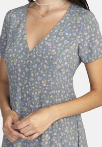 Kleid mit blauen Blumen, V-Ausschnitt, kurzen Ärmeln und Knöpfen an der Vorderseite. Der Stoff hat kleine rosa und grüne Blumenmuster.
