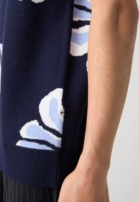 Marineblauer Strickpullover mit abstrakten hellblauen und weißen Mustern, mit gerippten Bündchen und einem Etikett an der Seitennaht.