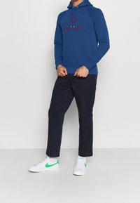 Sudadera azul con logo rojo "SAINT", bolsillo canguro y capucha con cordón, combinada con pantalones oscuros y zapatillas blancas con acentos verdes.