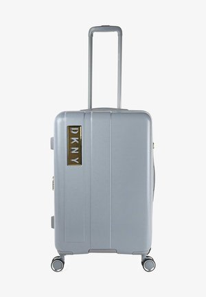 Maleta rígida plateada con surcos verticales, mango retráctil y cuatro ruedas. Logo de DKNY en dorado en la parte frontal.