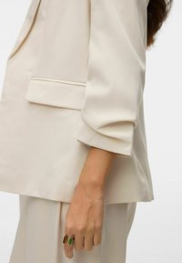Beżowa, dopasowana marynarka z podwiniętymi rękawami, z widoczną kieszenią i gładką fakturą. Ręka ozdobiona jest dwukolorowym pierścieniem.