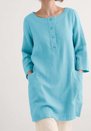 Jasnoniebieska lniana tunika z długimi rękawami, okrągłym dekoltem i plisą na guziki. Posiada dwie przednie kieszenie o prostym kroju.