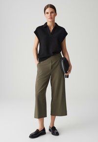 Top corto nero texturizzato, pantaloni larghi a vita alta verde oliva e mocassini neri. Gli accessori includono una piccola borsa nera.