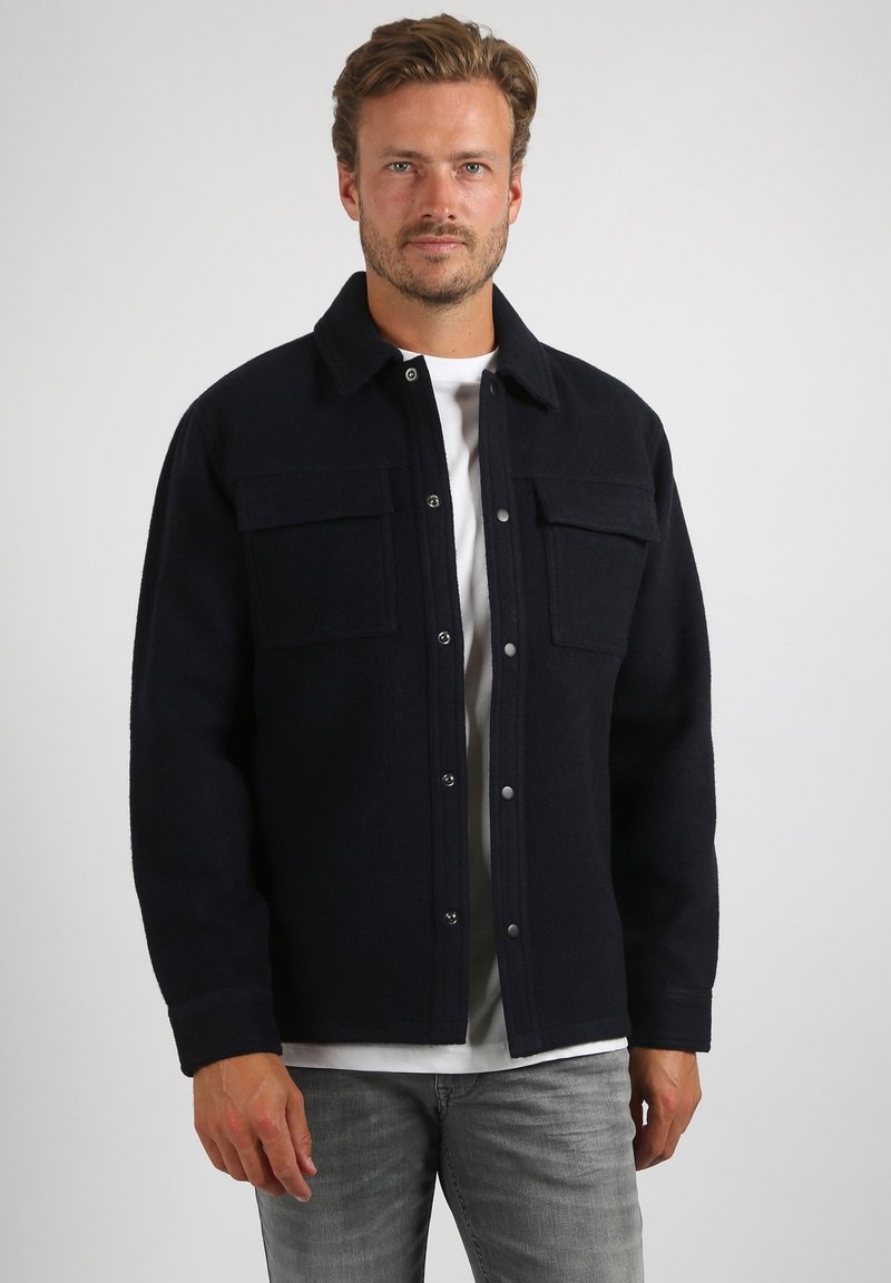 Man in een zwarte button-up jasje met borstzakken over een wit shirt en grijze jeans, staande tegen een effen lichte achtergrond.