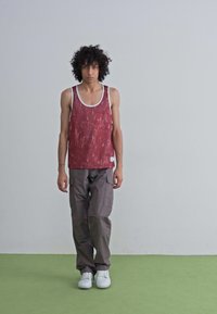 Débardeur à motif rouge avec des bordures blanches associé à un pantalon cargo gris. Le modèle porte des baskets blanches sur un sol vert.
