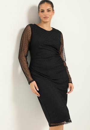 Femme appuyée contre un mur, portant une robe noire ajustée avec des manches longues transparentes et un motif discret à pois.