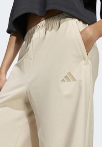 Béžové sportovní kalhoty s elastickým pasem, bočními kapsami a prořezávaným logem se třemi pruhy na stehně. Měkká, hladká látka.