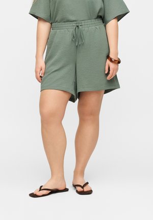 Ember zöld, texturált rövidnadrágot visel gumis derékpánttal és zsinórral, barna flip-flop papucsokkal és barna karkötővel a csuklóján.