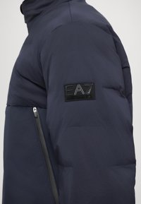 Jachetă pufosă de culoare bleumarin, cu un patch elegant EA7 de culoare neagră pe mânecă și o fantă cu fermoar de contrast pe lateral.