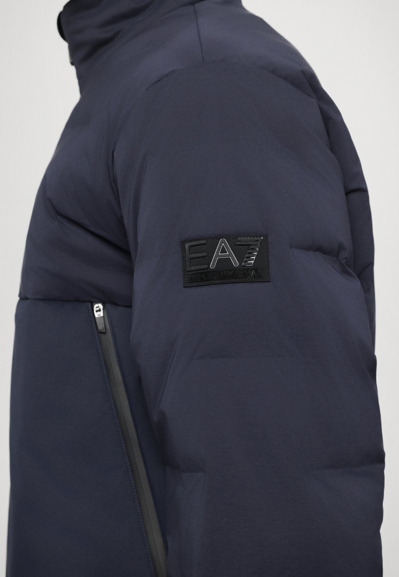 Marineblaue, gefütterte Jacke mit einem erhabenem schwarzen EA7-Logo-Patch an den Ärmeln und einer kontrastierenden, mit Reißverschluss versehenen Tasche an der Seite.