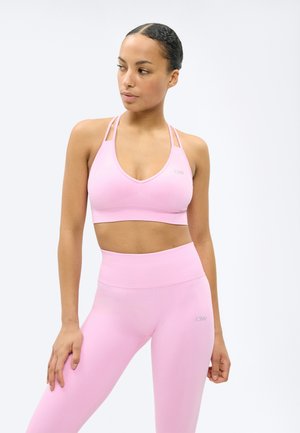 Γυναίκα που φοράει ανοιχτό ροζ αθλητικό σουτιέν και ψηλόμεσο κολάν, στέκεται με το ένα χέρι στη μέση, κοιτάζοντας προς τα πλάγια μπροστά σε λευκό φόντο.