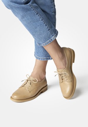 Beige leather oxford shoes with round toes, lace-up closure, and a smooth texture, paired with rolled-up blue denim jeans.