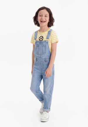 Enfant souriant avec des cheveux courts bouclés portant un t-shirt à rayures jaunes, une salopette en denim bleu clair et des baskets blanches, debout sur un fond blanc.