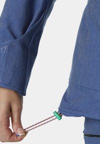 Blaue Jeansjacke mit einer Hand, die einen mehrfarbigen Kordelzug am Saum anpasst. Der Stoff wirkt strukturiert und hat ein lässiges Design.