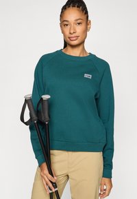 Žalsvos spalvos džemperis su raglano rankovėmis, ribinėmis rankogalais ir apačia. Yra mažas logotipo lopas. Du žygių lazdos su juodais rankenomis.