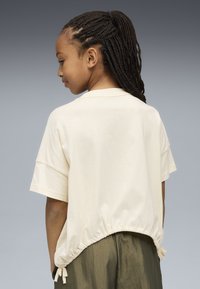 Joven con cabello trenzado que lleva una camiseta suelta de color crema con mangas cortas y cordones, y pantalones verde oliva, sobre un fondo gris.
