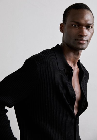 En svart man i en delvis knäppt svart ribbad kofta poserar mot en neutral ljus bakgrund och ser självsäkert in i kameran.