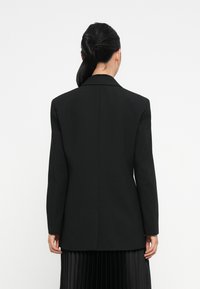 Blazer nero sartoriale con maniche lunghe, colletto a revers e design dritto sul retro. Texture liscia, dettagli minimi e vestibilità strutturata.