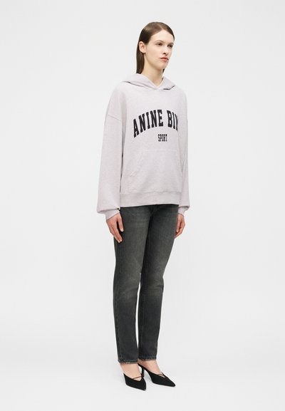 Woman standing wearing a light gray oversized hoodie with "ANINE BIR" text, dark gray jeans, and black pointed high heels.