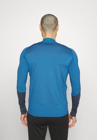 Maglietta sportiva a maniche lunghe di un vivace blu, con maniche nere texturate e un motivo geometrico. Design liscio e aderente.