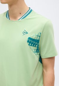 Személy világoszöld rövid ujjú inget visel, mintás türkiz betéttel és türkiz-fehér csíkos gallérdísszel.