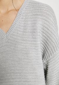Maglione grigio chiaro lavorato a maglia con scollo a V, caratterizzato da un motivo a punto testurizzato e maniche ampie.