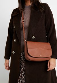 Hnědá kožená crossbody taška s klopou, se zlatým logem a nastavitelným popruhem, v kombinaci s tmavým kabátem a vzorovanou sukní.