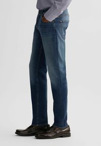 Jeans in denim blu scuro con vestibilità dritta; presenta un classico design a cinque tasche e dettagli di cucitura; indossati con scarpe eleganti marroni.