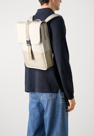 Mochila impermeable beige con un diseño elegante, que cuenta con un cierre superior y correas ajustables, llevada sobre una camisa de manga larga oscura.