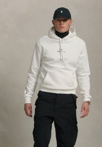 Sweat à capuche blanc avec poche frontale, présentant une petite broderie de logo. Un pantalon cargo noir et une casquette assortie complètent l'ensemble.