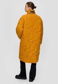 Prešívaný dlhý kabát v horčicovo žltej farbe, s vysokým golierom, geometrickou textúrou a zelenou visačkou na chrbte. Skombinovaný s čiernymi ribbed nohavicami.