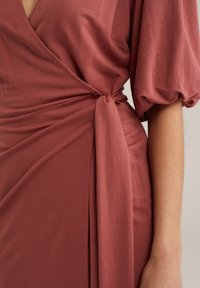 Zamatové šaty v rustikálnej farbe z mäkkej tkaniny; majú vypchaté rukávy a viazanie v pase, čo vytvára elegantný siluetu.