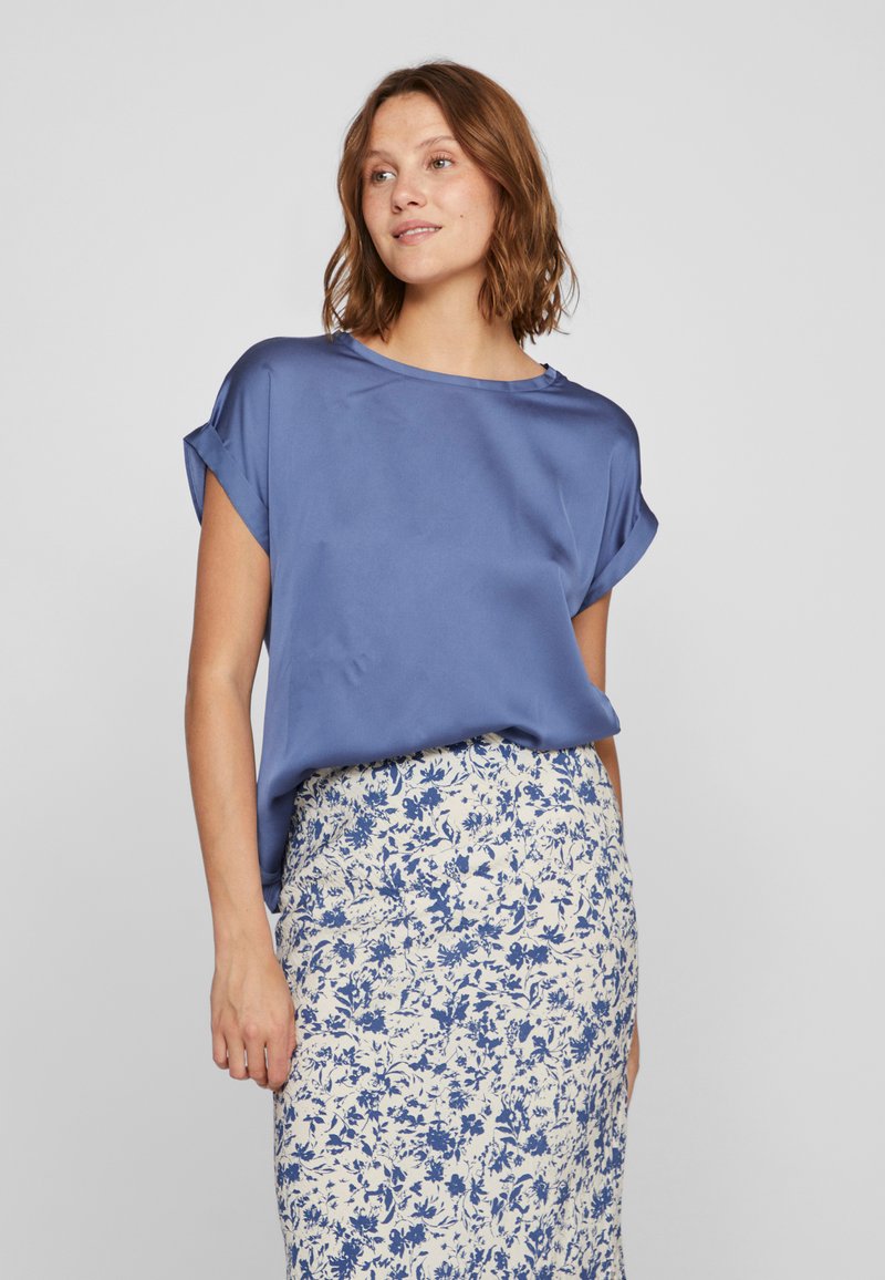 Blå silkebluse med korte oprullede ærmer; præsenteret over en blomster mønstret, cremet nederdel med blå accenter og en tætsiddende pasform.