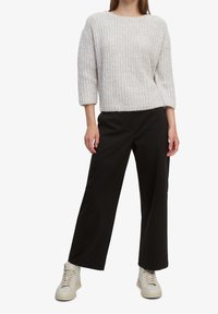 Chunky, lichtgrijze gebreide trui met 3/4 mouwen, gecombineerd met losse zwarte broeken met een zijzak. Off-white hoge sneakers.