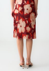 Gonna plissettata rossa e crema con motivo floreale, in tessuto texturizzato, design midi, caratterizzata da una vita aderente e una lucentezza sottile. Abbinata a décolleté in color nudo.