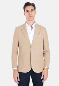 Blazer beige con texture, due tasche frontali, chiusura a singolo bottone e logo con accentuazione rossa. Indossato sopra una camicia bianca con un fazzoletto da taschino a fantasia.