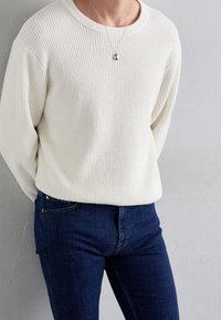 Kremowy prążkowany sweter z dzianiny z okrągłym dekoltem, zestawiony z ciemnoniebieskimi jeansami. Uzupełniony prostym srebrnym naszyjnikiem z zawieszką.
