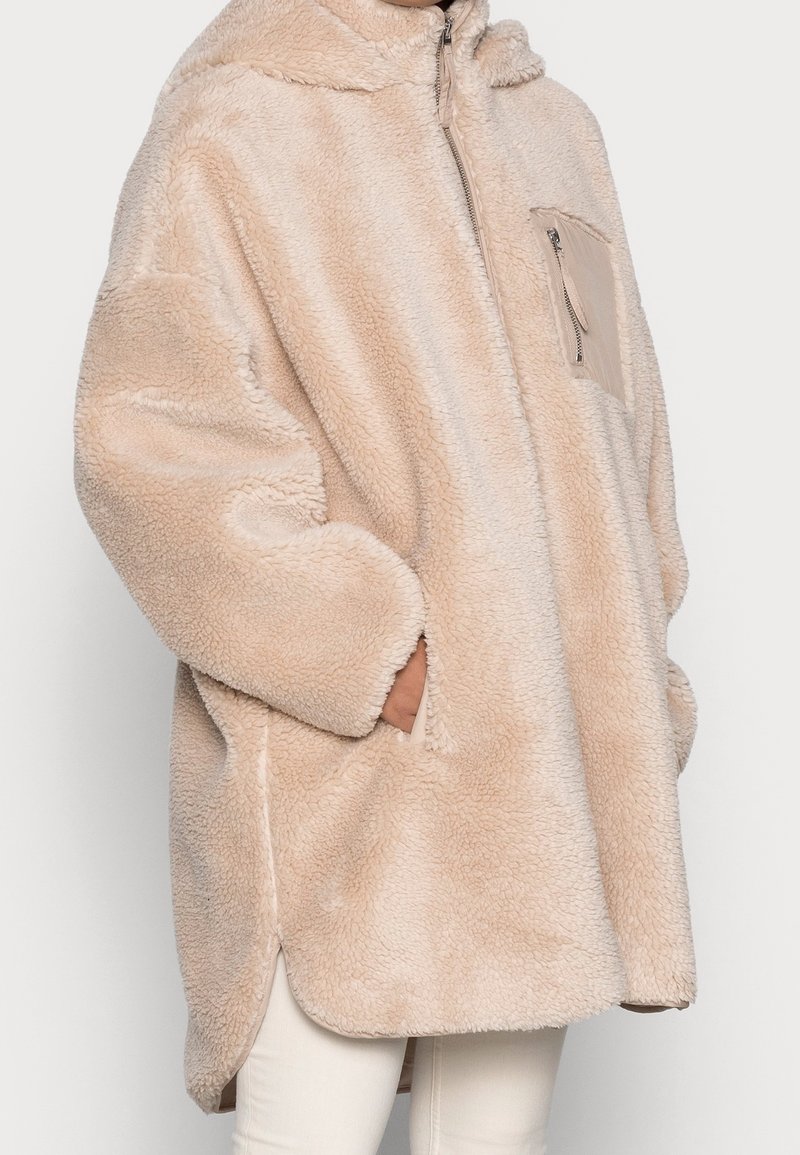 Beige, blød, pelslignende frakke med hætte, der har lynlås og en brystlomme. Blødt, tekstureret stof med sidelommer og løs pasform.