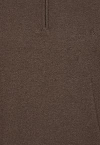 Ruskea neulepaita, jossa on lyhyt vetskari kaulassa ja pieni brodeerattu "X"-tyyli rinnassa.