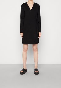 Svart omlottklänning med långa ärmar, V-ringning och sidoknytning. Bärs med svarta plattformssandaler. Slät textur, knälängd.