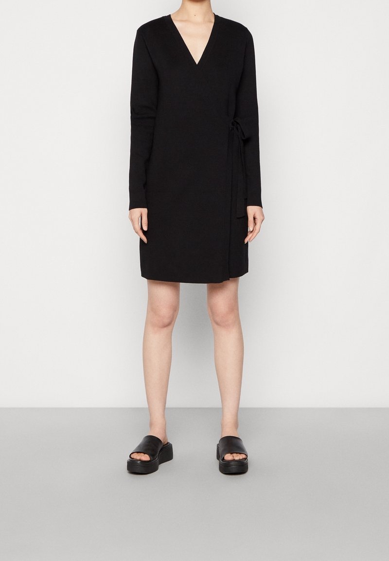 Svart omlottklänning med långa ärmar, V-ringning och sidoknytning. Bärs med svarta plattformssandaler. Slät textur, knälängd.