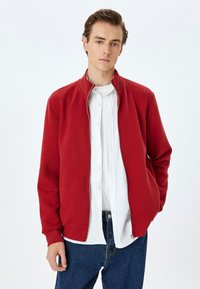 Giacca rossa a zip realizzata in tessuto liscio, con colletto alto e polsini a costine. Indossata sopra una camicia bianca con bottoni e jeans blu.