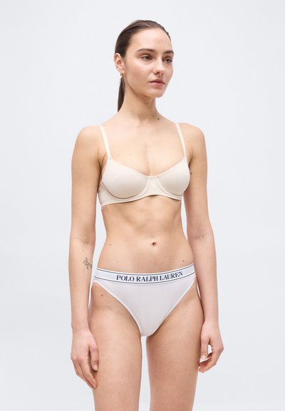 Γυναίκα που φοράει μπεζ σουτιέν με μπανέλα και λευκά εσώρουχα ψηλής κοπής με μαύρη μέση με λογότυπο "Polo Ralph Lauren", στέκεται μπροστά σε απλό φωτεινό φόντο.