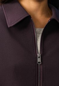Lilla zip-up jakke med krave. Har en sølv lynlås og blød tekstur, båret over en lysegrå top. Nærbillede af stoffet og lynlåsen.